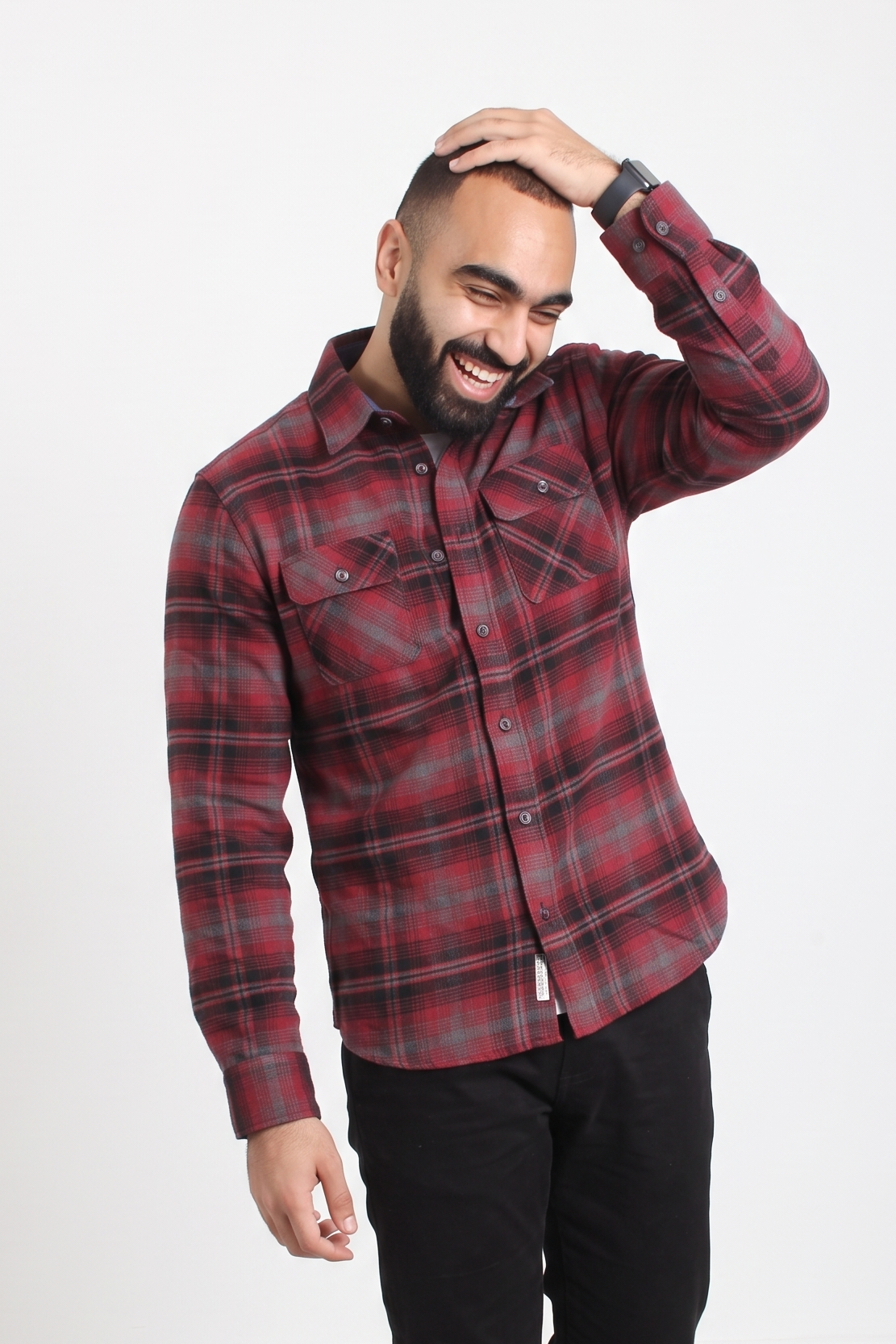 A man wearing a red and black plaid button-up flannel shirt and black pants, standing against a plain white studio background.