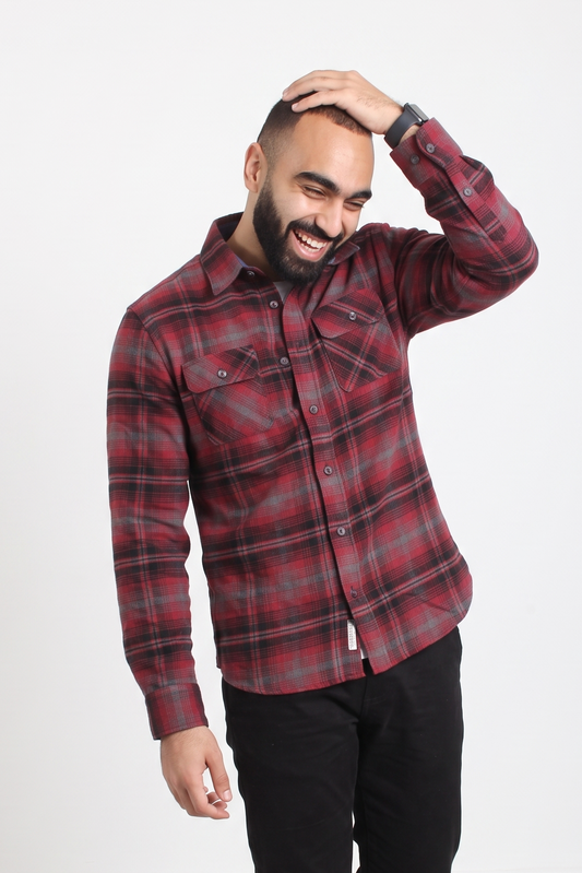A man wearing a red and black plaid button-up flannel shirt and black pants, standing against a plain white studio background.