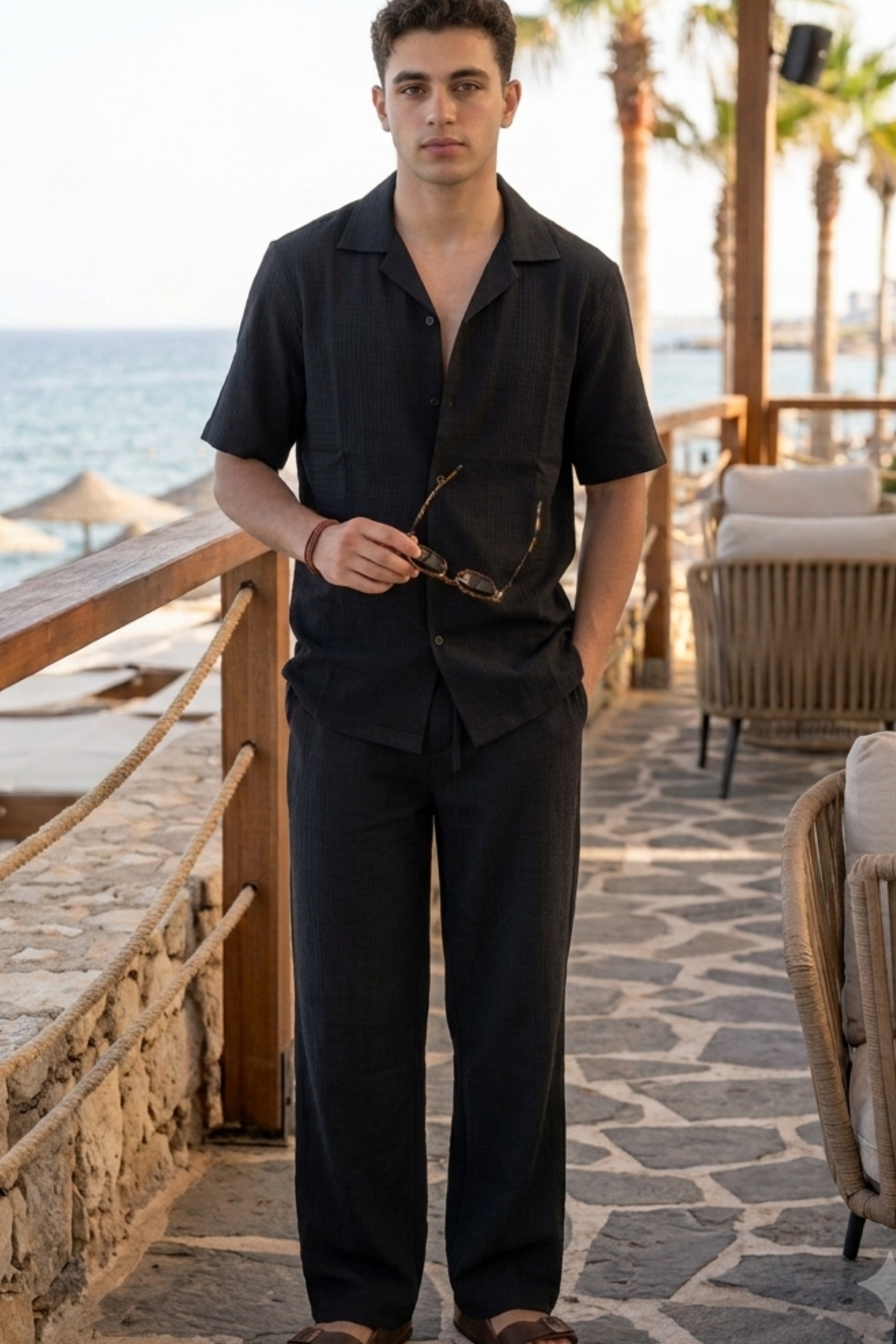 A man wearing a black short-sleeve button-up shirt and matching black trousers, standing on a stone patio with a beach and lounge chairs in the background.