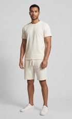 A man modeling a matching cream-colored lounge set consisting of a short-sleeve crewneck t-shirt and drawstring shorts, paired with white sneakers, against a light gray studio background.