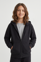 A young girl with wavy brown hair smiling, wearing a black zip-up hoodie over a light gray t-shirt, photographed against a solid light gray background.