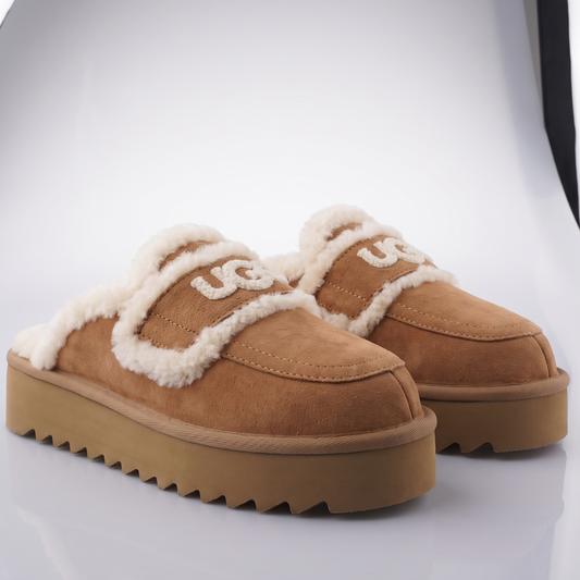 A pair of UGG slip-on mule slippers in tan suede with a cream shearling lining and a chunky lug sole, displayed on a reflective white surface.